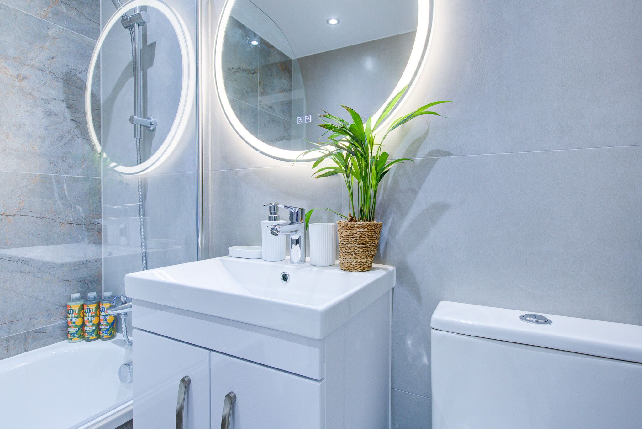 A modern bathroom with a white sink, two round mirrors, a shower, and a toilet.