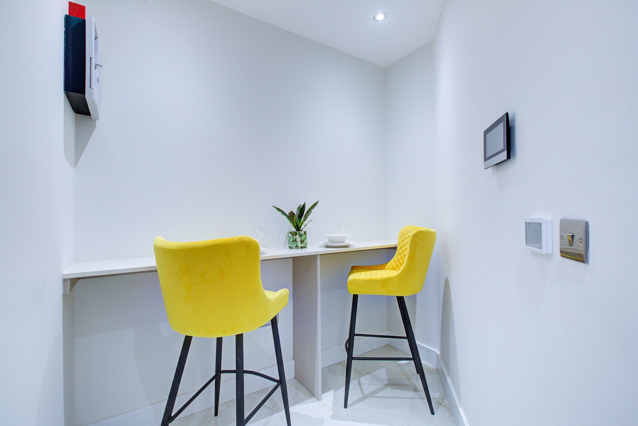 Une petite pièce moderne avec deux chaises jaunes devant un bureau blanc, une petite plante en pot et un luminaire fixé au mur.
