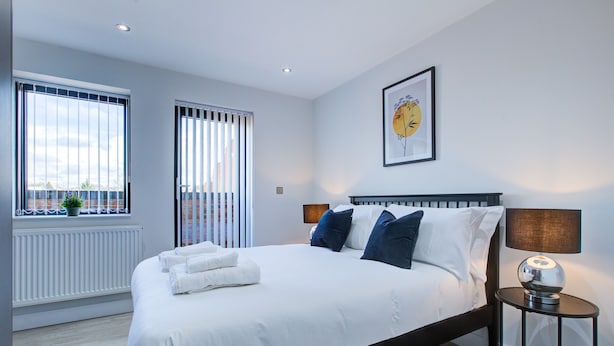 A modern bedroom with a large bed, a side table, and a window with blinds. - Hybrid Resi - Uxbridge  (Uxbridge)