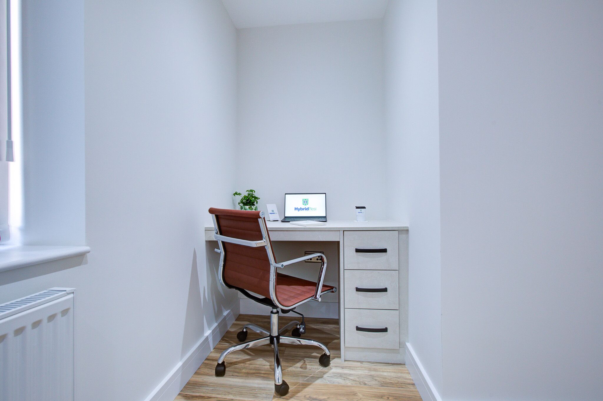 A corner office with a white desk, a brown office chair, a small plant, and a sign reading "Hybrid."