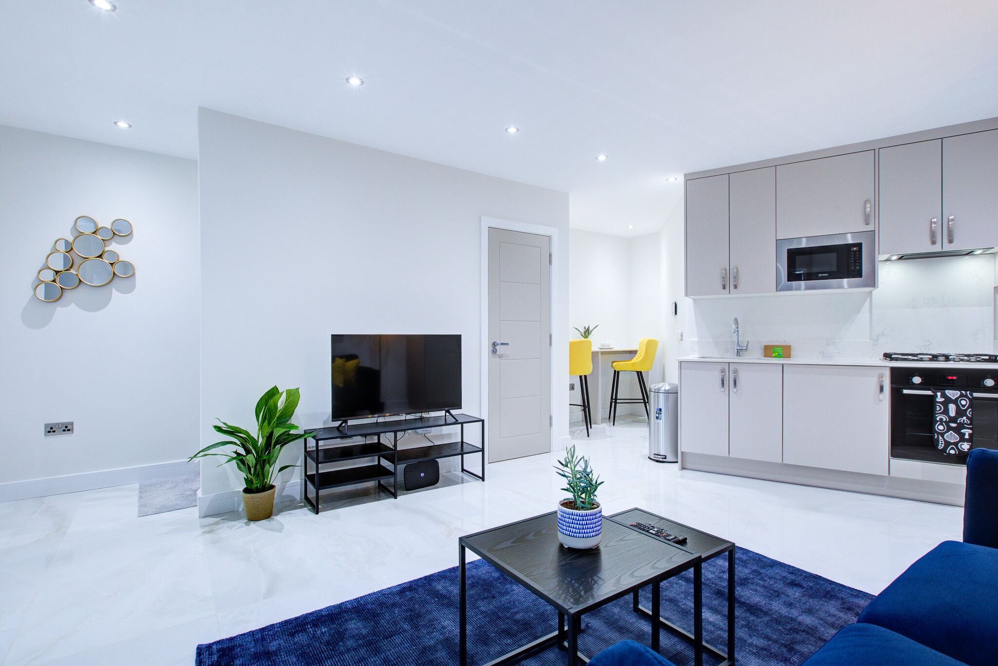 A modern living room with a flat-screen TV, a coffee table, and a blue sofa. There is a kitchen area with grey cabinets and a microwave.