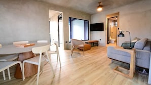 Ein modernes Wohnzimmer mit Holzboden, einer grauen Couch, einem Esstisch mit Stühlen und einem an der Wand montierten Fernseher.