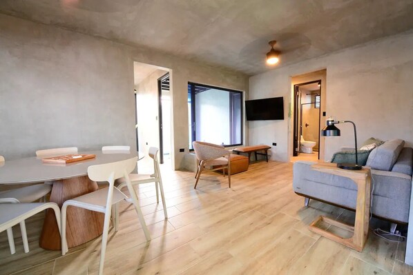 A modern living room with a wooden floor, a grey sofa, a dining table with chairs, and a TV mounted on the wall.