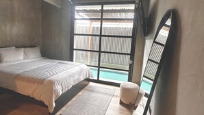 Ein Schlafzimmer mit Bett, einem kleinen Hocker, einem Spiegel und Blick auf einen Swimmingpool durch ein großes Fenster.