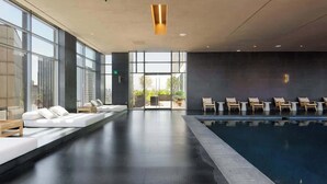 Un espace piscine intérieur moderne, doté de chaises longues, d’un coin salon avec des coussins et de grandes fenêtres.