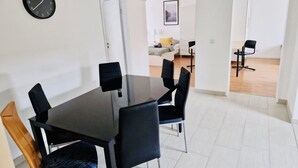Un espace repas moderne avec une table et des chaises noires, une horloge au mur et une porte blanche donnant sur une autre pièce.