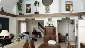 A spacious living area with a high ceiling, wooden beams, and a dining table with chairs. There are several pieces of artwork on the walls, a staircase leading to another level, and a variety of furniture including a sofa, chairs, and a lamp.