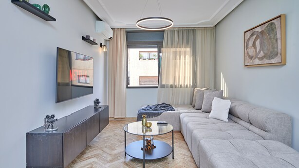 Una sala de estar moderna con un televisor de pantalla plana, una mesa de centro de vidrio, un sofá gris y un ventanal con cortinas.