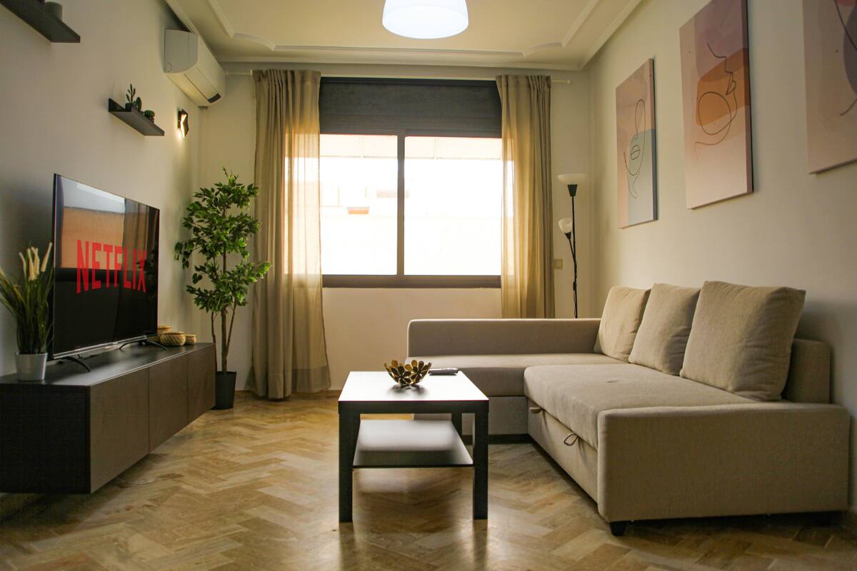 A living room with a flat-screen TV, a sofa, a coffee table, a potted plant, and a window with curtains.