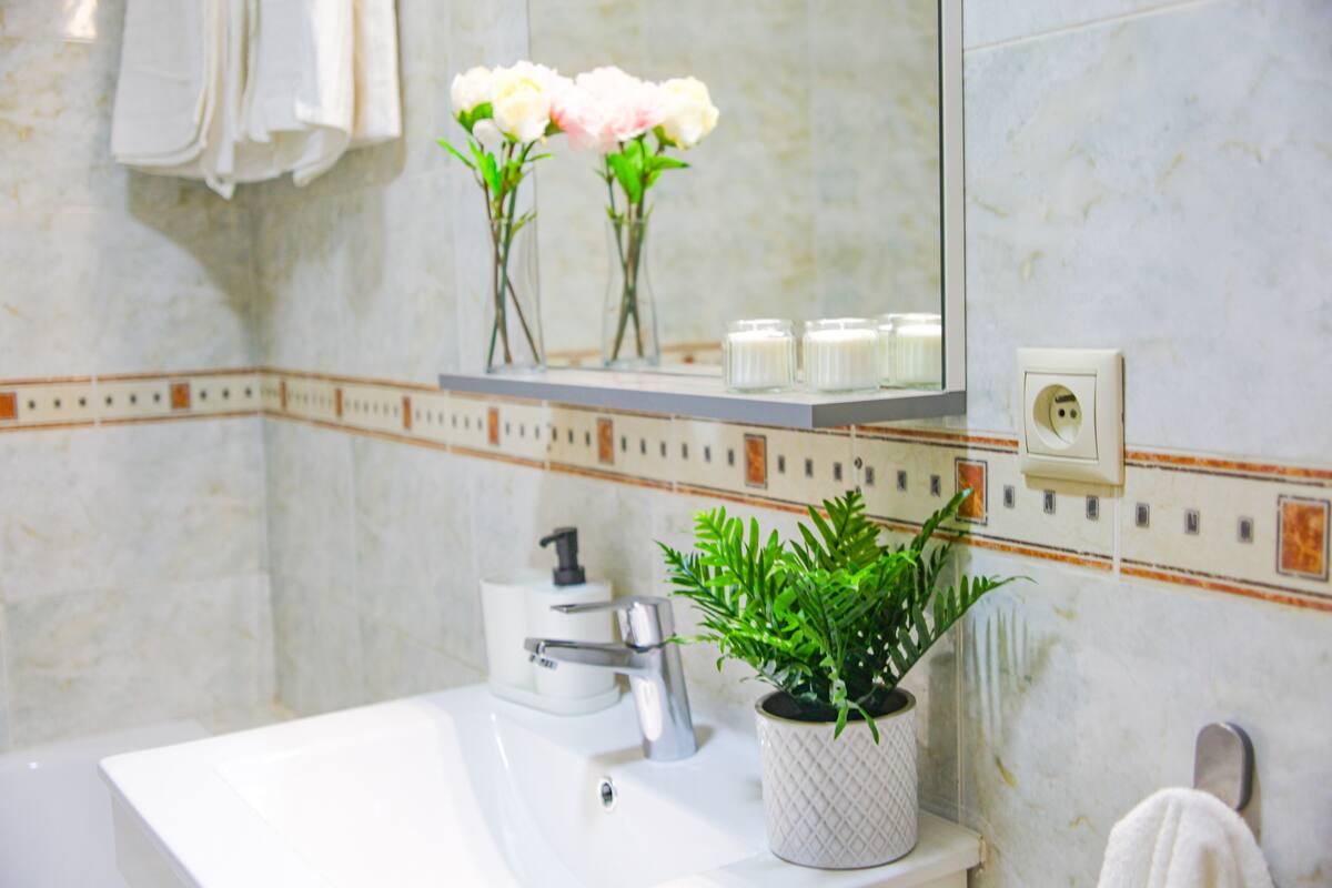 A bathroom with a white sink, a mirror, a potted plant, and a shelf with flowers.