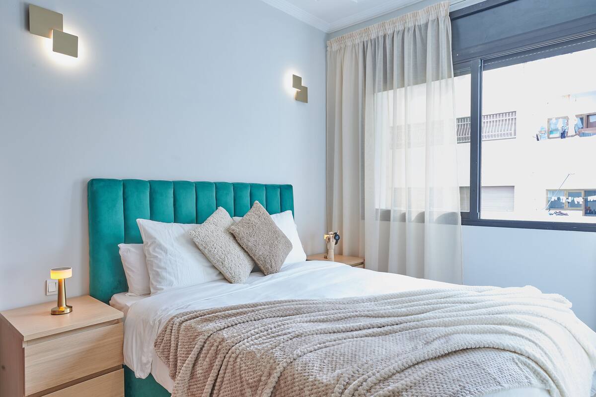 A modern bedroom with a teal headboard, white bedding, and a view of the city through a large window.
