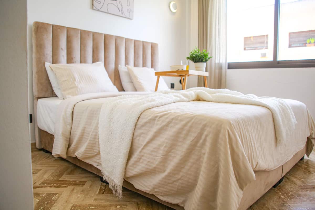 A neatly made bed with a headboard, a bedside table with a plant, and a window with curtains.