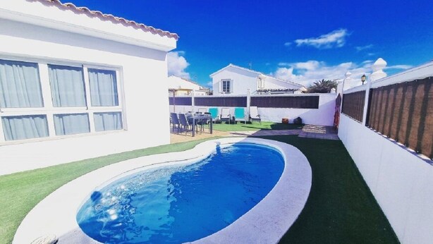 A swimming pool surrounded by a white house with a tiled roof, a small garden, and a clear blue sky.