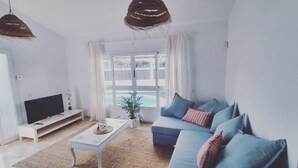 A modern living room with a flat-screen TV, a sofa with blue cushions, a coffee table, and a large window with a view of a pool.