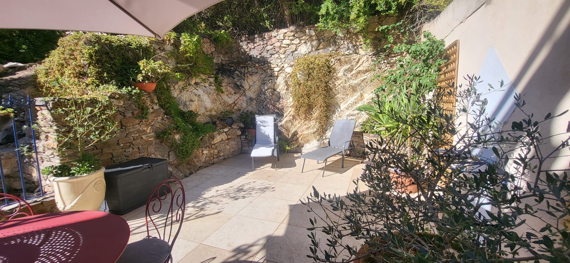 An outdoor patio with stone walls, two white lounge chairs, a red chair, and potted plants.