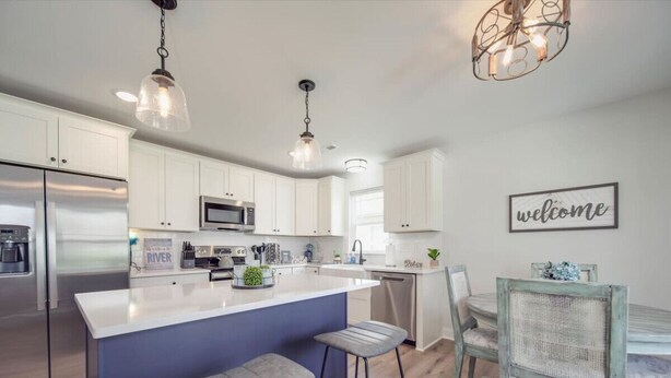 A modern kitchen with white cabinets, a central island with stools, stainless steel appliances, and a 'welcome' sign on the wall.