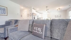 A modern living room with a grey sofa, a 'STAY awhile' pillow, and a 'welcome' sign on the wall.