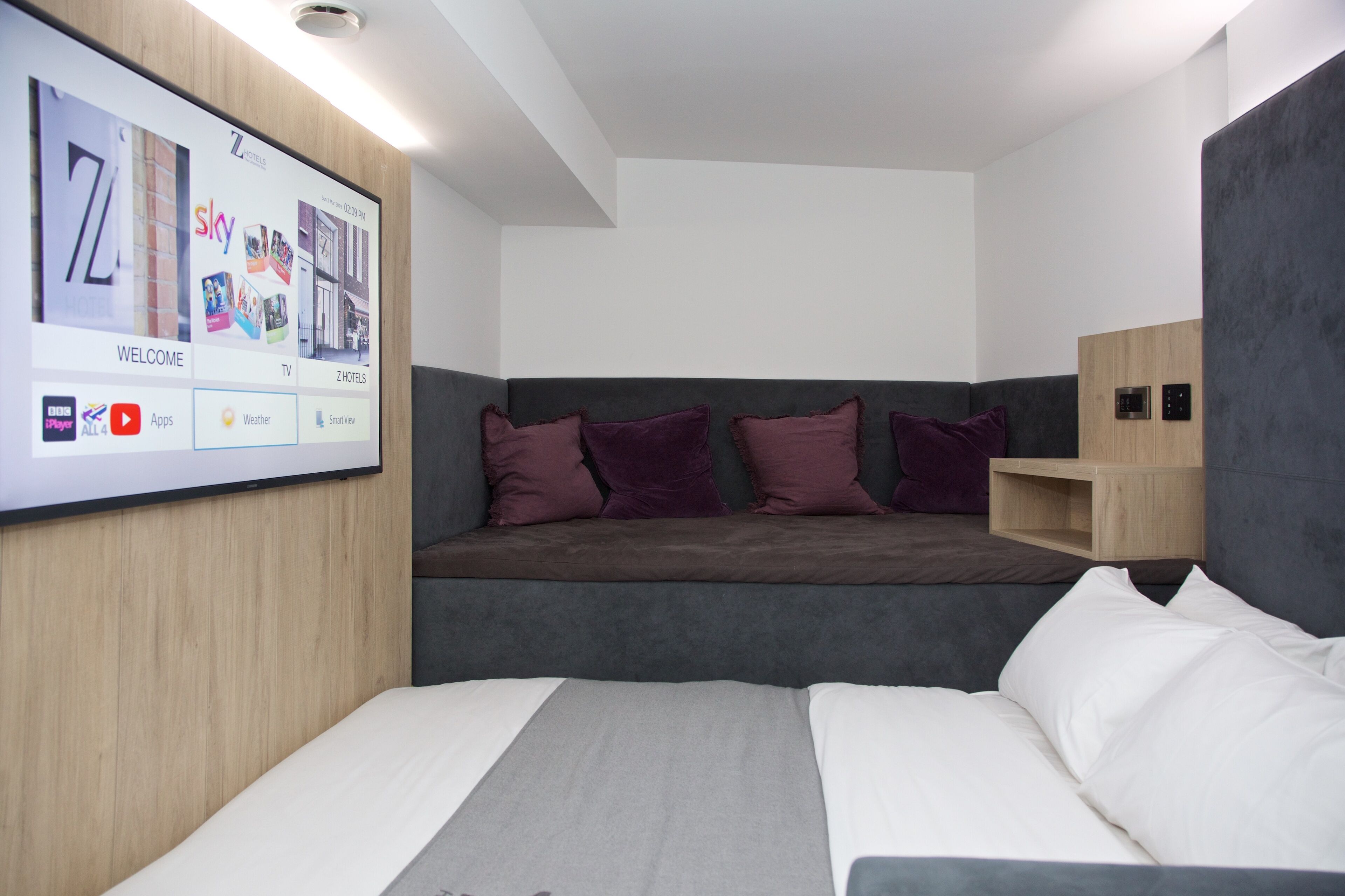 A modern hotel room with a large TV screen displaying a welcome screen, a grey sofa with purple cushions, a wooden shelf unit, and a bed with white bedding.