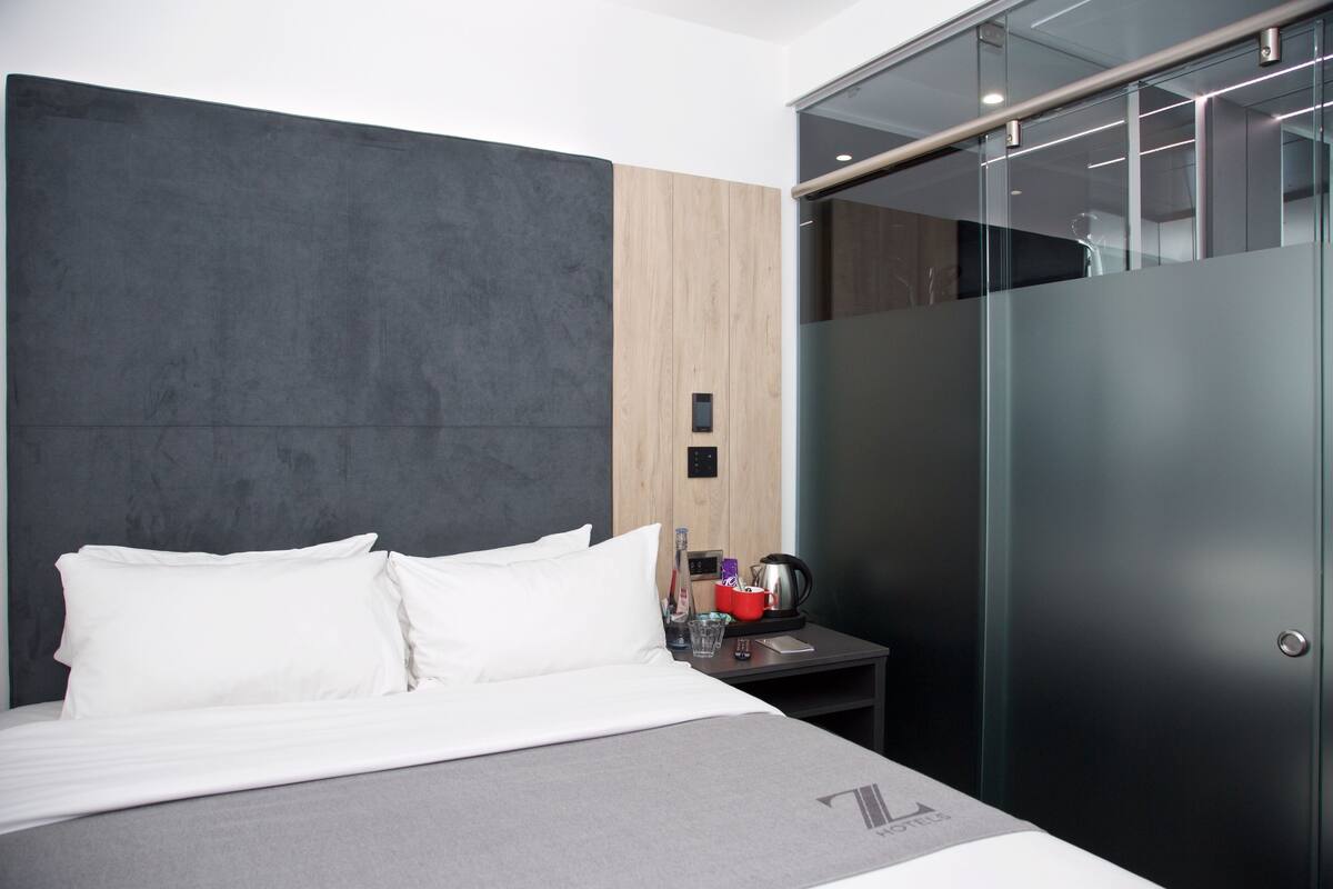 A modern hotel room with a large bed, a bedside table with a red mug, a glass of water, and a coffee maker.