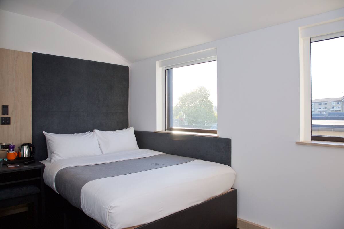 A modern hotel room with a large bed, a bedside table with a kettle, and a window with a view of buildings.