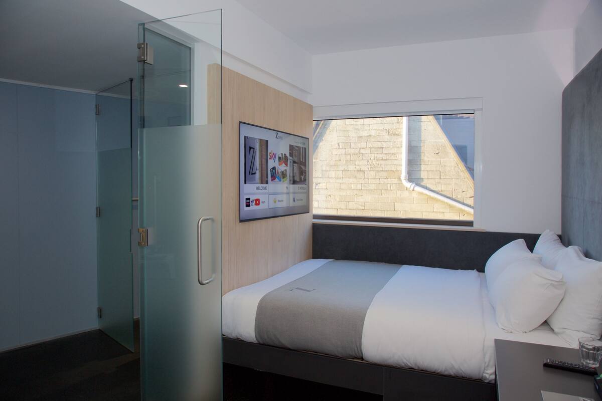 A modern hotel room with a bed, a glass shower enclosure, a mounted television, and a window with a view of the outside.