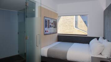 Une chambre d’hôtel moderne équipée d’un lit, d’une douche à parois de verre, d’un téléviseur fixé au mur et d’une fenêtre donnant sur l’extérieur.