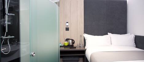 A modern hotel room with a glass shower enclosure, a bedside table with a kettle, and a bed with white linens.