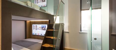 A compact room with a bunk bed, a TV mounted on the wall, a glass staircase, and a door with a frosted glass panel.