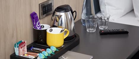 Quarto de hotel com cafeteira, caneca amarela, controle remoto e bandeja com sachês de chá e copos.