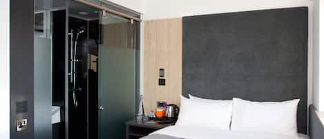 Ein modernes Hotelzimmer mit einem großen Bett, einem gläsernen begehbaren Kleiderschrank, einem Nachttisch mit Kaffeemaschine und einem kleinen Waschbecken.