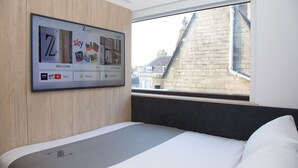 Une chambre d’hôtel dotée d’un grand écran numérique au mur, d’un lit avec une literie blanche et grise, et d’une fenêtre donnant sur un immeuble en briques.