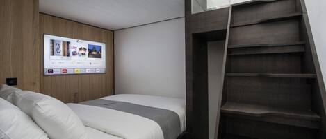 A modern hotel room with a bed, a mounted TV screen displaying a welcome message, and a built-in ladder leading to a mezzanine.