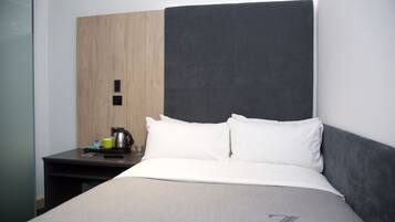 Una habitación de hotel con cama, mesita de noche, cafetera y vaso de agua.