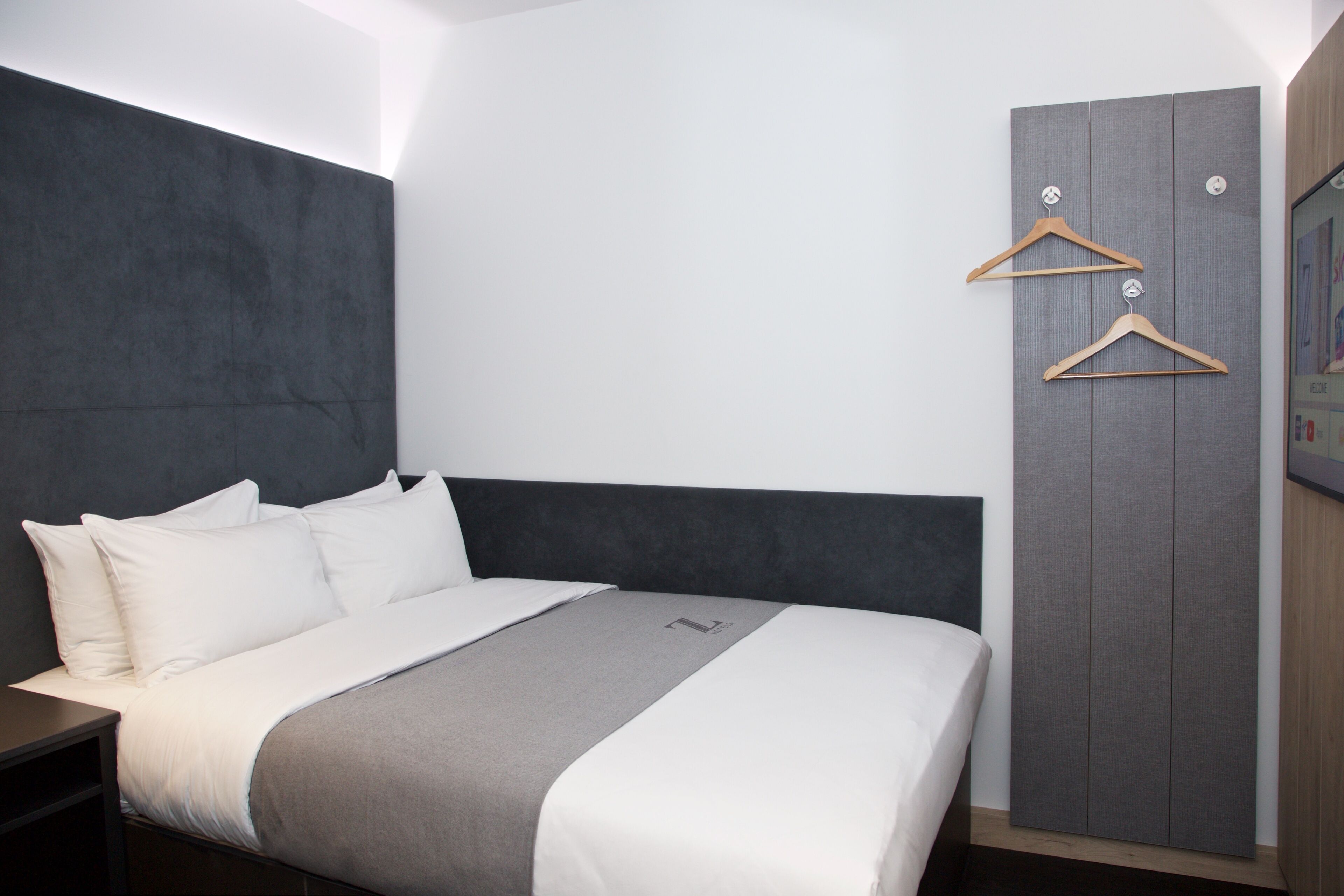Habitació d'hotel moderna amb un llit gran, un panell de capçalera gris, un armari amb penjadors de fusta i una televisió de pantalla plana.