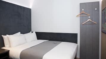 Habitació d'hotel moderna amb un llit gran, un panell de capçalera gris, un armari amb penjadors de fusta i una televisió de pantalla plana.