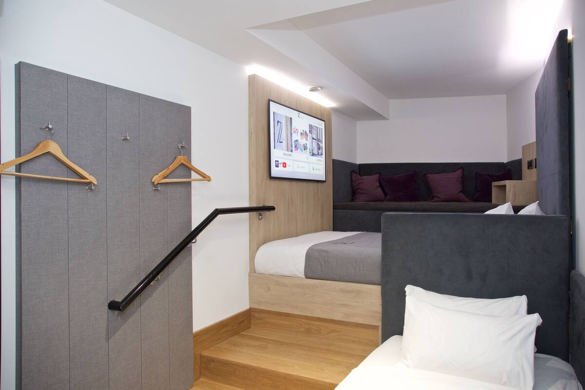 A modern hotel room with a grey wardrobe, a bed with white bedding, a grey headboard, and a TV mounted on the wall displaying a welcome screen.