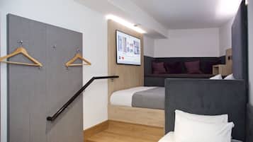 Habitación de hotel moderna con un armario gris, cama con ropa de cama blanca, cabecero gris y televisor montado en la pared mostrando una pantalla de bienvenida.