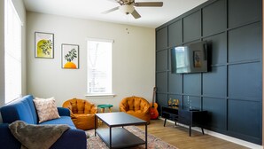 Ein Wohnzimmer mit einem blauen Sofa, orangefarbenen Sesseln, einem Couchtisch, einer Gitarre und einem an einer dunklen Wand befestigten Fernseher.