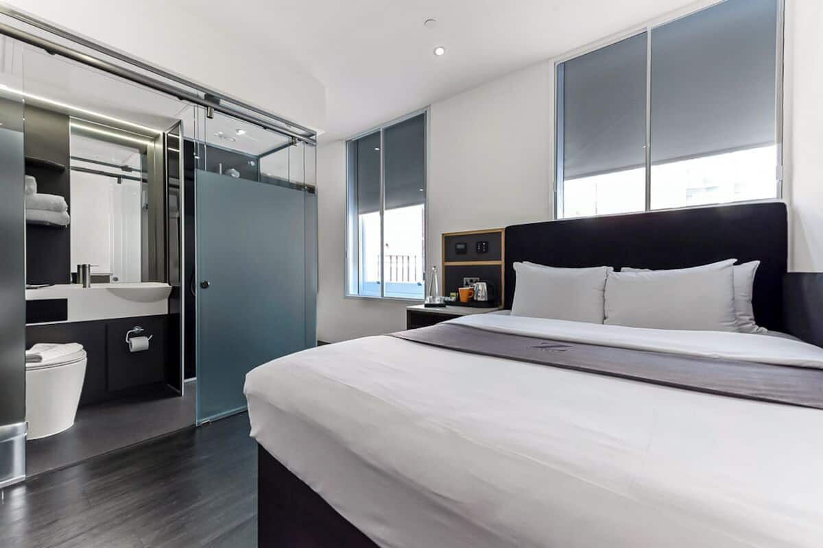 Moderni hotellihuone, jossa on suuri sänky, erillinen kylpyhuone lasiseinällä varustetulla suihkulla sekä ikkunasta avautuva näkymä kaupunkiin.