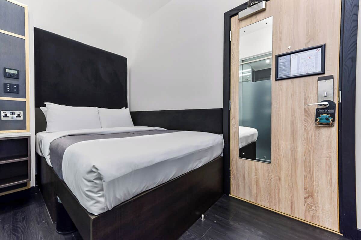 Moderni hotellihuone, jossa on suuri sänky, puuovi, pieni työpöytä tuolilla ja seinässä oleva ohjauspaneeli.
