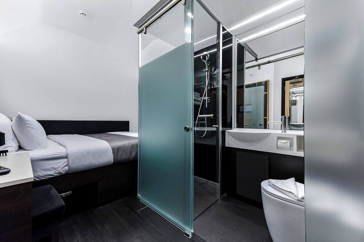 Moderni hotellihuone, jossa on sänky, lasiseinäinen suihkukaappi, pesuallas ja wc.