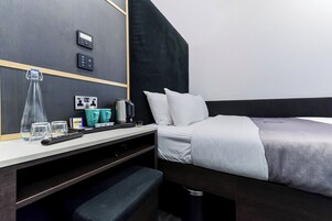 A hotel room with a bed, bedside table, and a small bench. There are control panels on the wall, a kettle, and a glass pitcher.