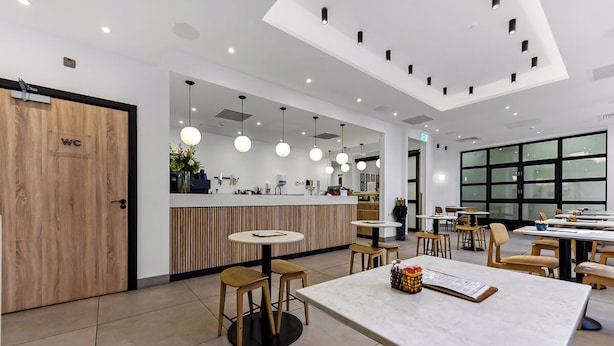A modern cafe interior with a wooden WC, round tables, hanging pendant lights, and a large window. - The Z Hotel Strand (London)