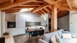 Cozinha moderna com armários brancos, sala de jantar com mesa e cadeiras de madeira, e sala de estar com sofá e almofadas.