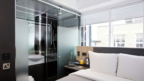A modern hotel room with a glass-enclosed shower, a bed with white bedding, a bedside table with a lamp, and a view of the city through large windows. - Z Tottenham Court Road (London)