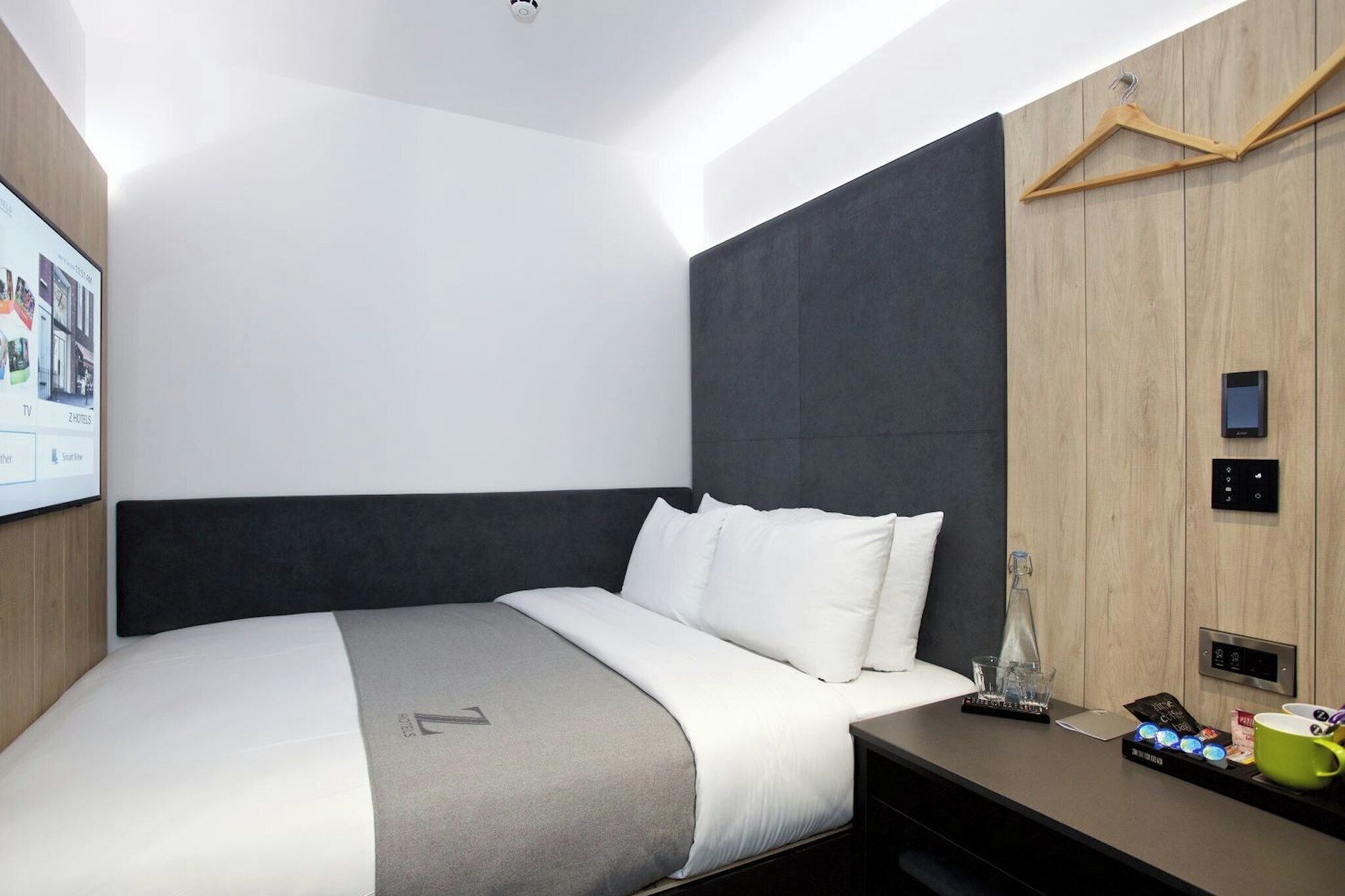 A modern hotel room with a large bed, a bedside table with a phone, a coffee maker, and a wall-mounted TV.