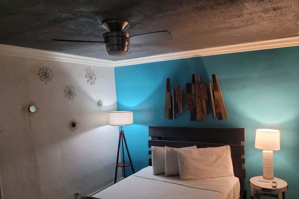 A bedroom with a bed, a ceiling fan, a nightstand with a lamp, and decorative wall art.