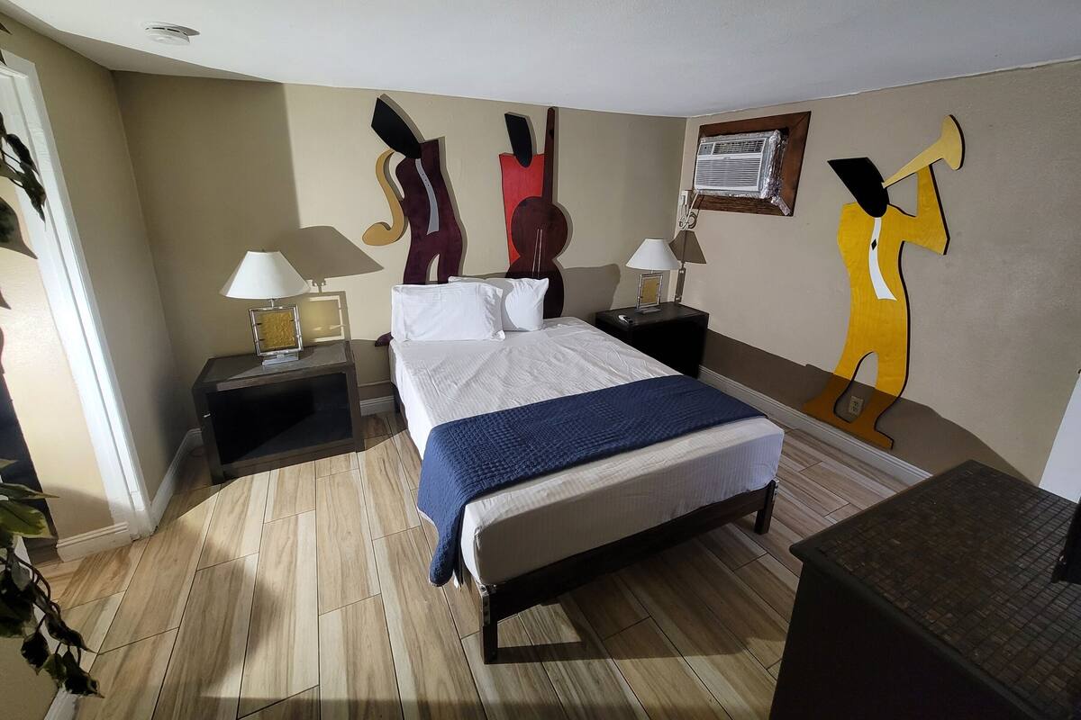 A hotel room with a bed, two bedside tables, a nightstand, a wall-mounted air conditioner, and a decorative wall piece.