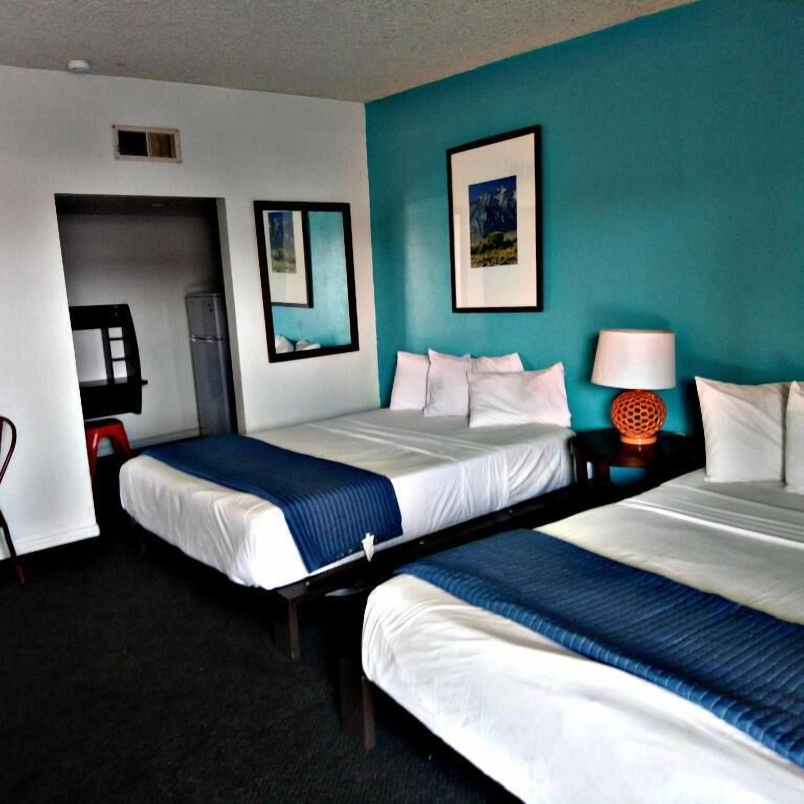 A hotel room with two beds, a teal accent wall, a framed picture, a lamp, and a small orange decorative item.