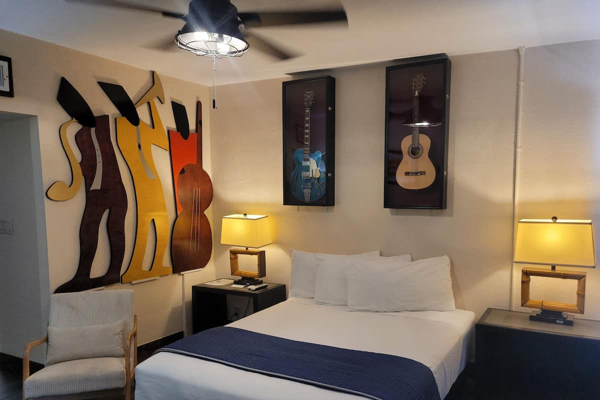 A hotel room with a bed, two nightstands, a chair, a ceiling fan, and wall art including a guitar and abstract figures.